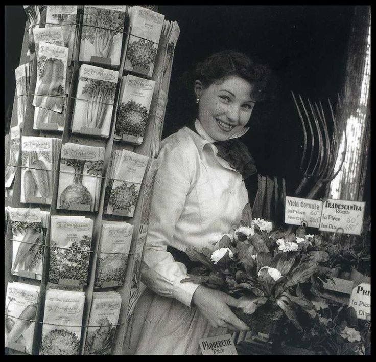 1930-tallet. Smilendeame som holder en blomst foran en hylle med frø. 